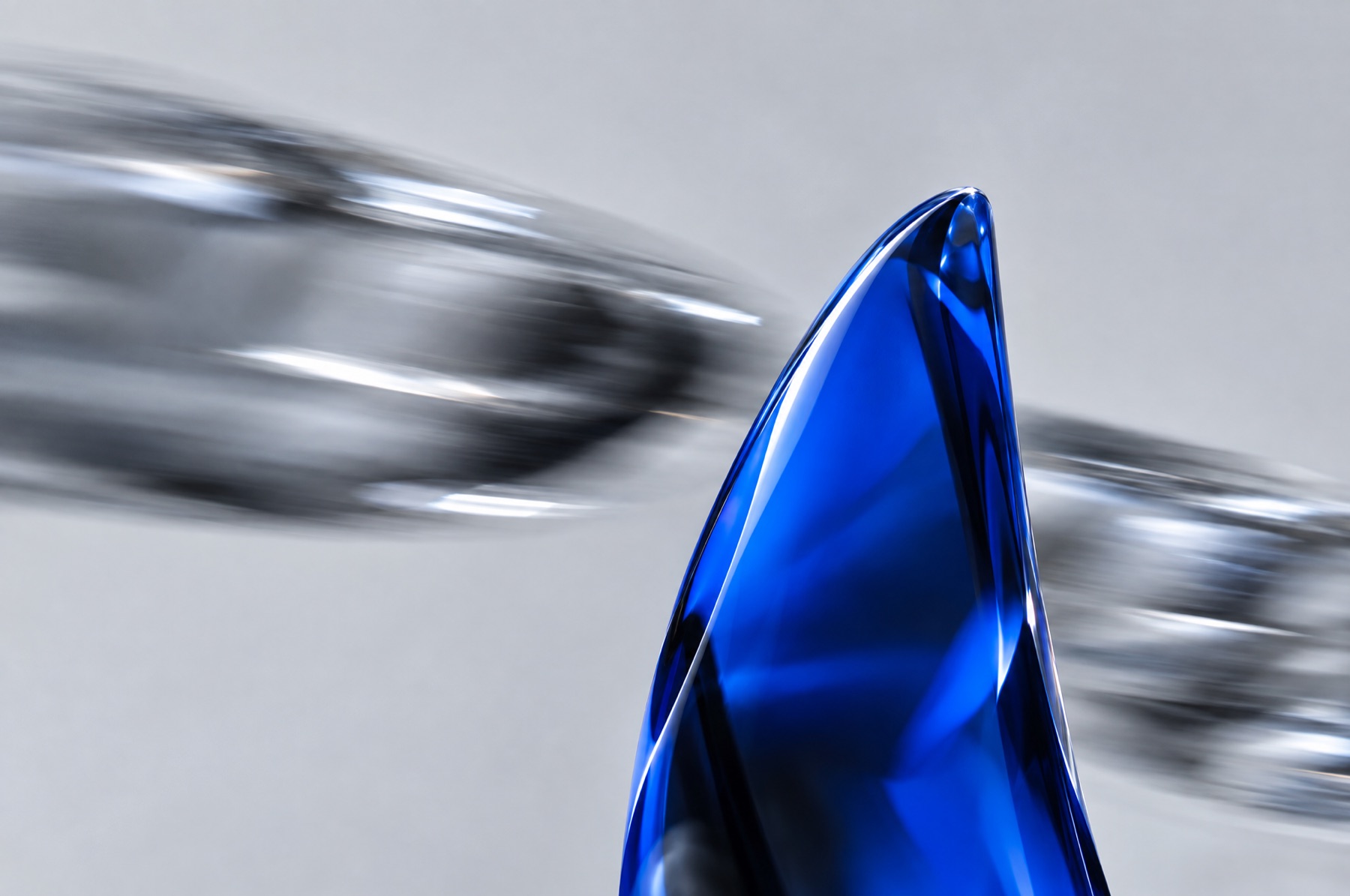 Eine scharf abgebildete blaue Glasform steht still, während verschwommene graue Glasformen im Hintergrund vorbeirasen, als Sinnbild für den Moment der Prüfung unter Zeitdruck.